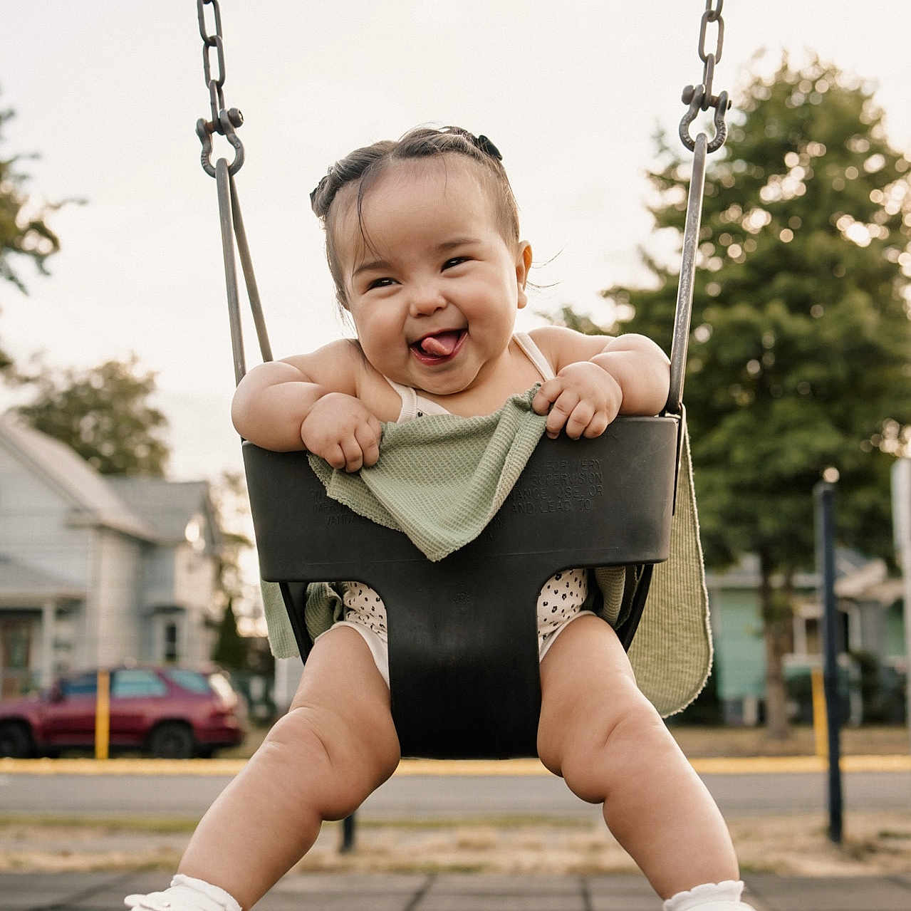 Jasmine is registered to the contest to win money with this photo: baby, chains, child, clothing, face, fun, green_blanket, happy, houses, legs, outdoor, person, playground, sandals, sidewalk, smiling, sunset_light, swing, tongue_out, trees