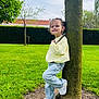 child, toddler, smiling, tree_trunk, stick, jeans, white_sneakers, yellow_sweater, green_grass, park, outdoor, nature, hedge, sky, pose, portrait, daylight, playful, young_child, happy
