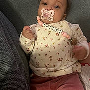 Kaysha participe au concours pour gagner de l'argent avec cette photo : baby, beaded_chain, calm, child, clothing, comfort, cute, face, floral_pattern, hand, indoor, infant, lying_down, name_beads, pacifier, person, pink_pants, portrait, sweater, textured_surface