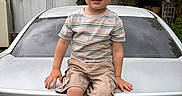 Liam is registered to the contest to win money with this photo: child, boy, car, dodge, trunk, striped_shirt, cargo_shorts, shoes, outdoor, nature, greenery, shed, smiling, casual, portrait, young, person, daylight, vehicle, happy
