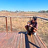 dog, doberman, person, outdoor, sunny, deck, wooden_floor, happiness, embrace, leash, nature, grassland, blue_sky, sunglasses, casual_clothing, smiling, pet, companionship, daytime, shadow