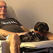 Beau joined the competition — help win amazing prizes! man, elderly, glasses, tshirt, dog, black_dog, scarf, armchair, indoor, relaxing, seated, pet, companion, calm, warm_lighting, furniture, home, casual, comfortable, resting