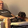 man, elderly, glasses, tshirt, dog, black_dog, scarf, armchair, indoor, relaxing, seated, pet, companion, calm, warm_lighting, furniture, home, casual, comfortable, resting