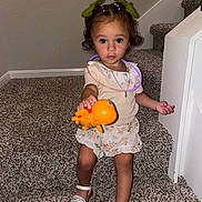 Mia is registered to the contest to win money with this photo: toddler, child, toy, orange_toy, stairs, carpet, green_bows, hair_accessories, necklace, glow_stick, sitting, curious, indoor, white_sandals, floral_skirt, short_sleeve_top, young_child, playful, looking_at_camera, home