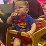 Zayden is registered to the contest to win money with this photo: baby, child, high_chair, superman, costume, blue_shirt, red_bow_tie, snack, eating, sitting, wooden_chair, indoor, event, crowd, portrait, face, smiling, infant, superhero, warning_label