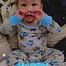 baby, child, sitting, smiling, happy, birthday, confetti, text_overlay, pajamas, onesie, blue_clothing, colorful_clothing, indoors, couch, cushions, portrait, playful, hands_on_face, barefoot, celebration