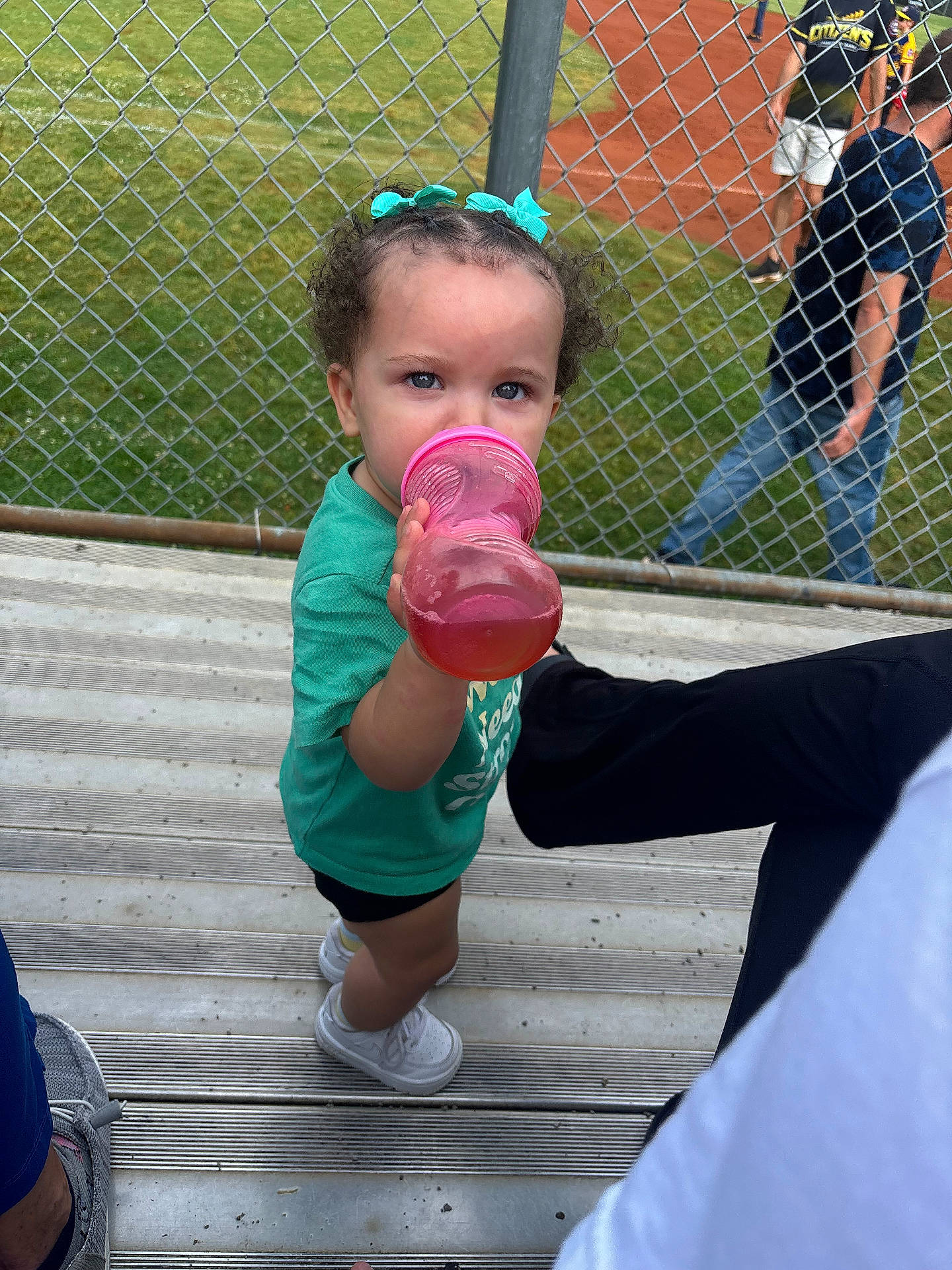 Nyah joined the competition — help win amazing prizes! baseball_field, bleachers, casual_clothing, chain_link_fence, child, cup, curly_hair, daylight, drinking, grass, green_shirt, hair_bows, outdoor, person, playful, spectator, sports, toddler, white_shoes, young_child
