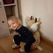 Calista a rejoint le concours — aidez-le/la à gagner de superbes lots ! child, toddler, plush_toy, stuffed_animal, reindeer, smile, blonde_hair, pajamas, slippers, indoor, wooden_floor, shelf, cabinet, wall, corner, cozy, portrait, candid, toy_face, furniture