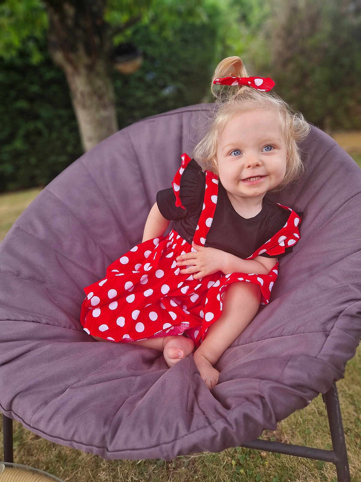 Calista participe au concours pour gagner de l'argent avec cette photo : baby, toddler, child, red_dress, polka_dots, hair_bow, blue_eyes, blond_hair, smile, happy, portrait, papasan_chair, cushion, outdoor, garden, grass, tree, barefoot, playful, rosy_cheeks