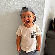 Josiah is registered to the contest to win money with this photo: toddler, child, boy, smiling, cap, tshirt, shorts, indoor, wall, floor, couch, bag, happy, standing, casual_clothing, portrait, cute, young_child, joyful, home
