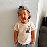 toddler, child, boy, smiling, cap, tshirt, shorts, indoor, wall, floor, couch, bag, happy, standing, casual_clothing, portrait, cute, young_child, joyful, home