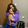 backpack, bedroom, blue_pants, cat, child, curly_hair, electrical_outlet, girl, hugging, indoor, kitten, mirror, orange_cat, person, pet, portrait, purple_jacket, smiling, string_lights, wooden_door
