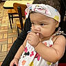 adult_holding_child, baby, cafe, cartoon_character, chairs, child, curious, dress, earrings, floral_pattern, hand_to_mouth, headscarf, indoor, infant, minnie_mouse, portrait, restaurant, sitting, tables, tile_floor