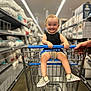 aisle, baby, black_shirt, checkered_shoes, child, clothing, floor, hand, happy, indoor, person, pillows, retail, shelves, shopping, shopping_cart, smiling, store, toddler, walmart