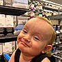 aisle, baby, bedding, black_clothing, boxes, child, cute, face, happy, head, indoor, mischievous, person, retail, shelf, shopping_cart, smile, store, toddler, young_child