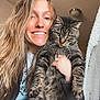 animal, casual_clothing, cat, cozy, expression, face, furniture, hand, happy, holding, home, indoor, jewelry, light, long_hair, pet, portrait, smiling, tabby_cat, woman