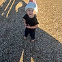 Kyle is registered to the contest to win money with this photo: toddler, child, barefoot, hat, pom_pom, black_clothing, outdoor, wood_chips, sunlight, shadow, daylight, playground, curious, person, young_child, standing, casual, winter_hat, expression, nature