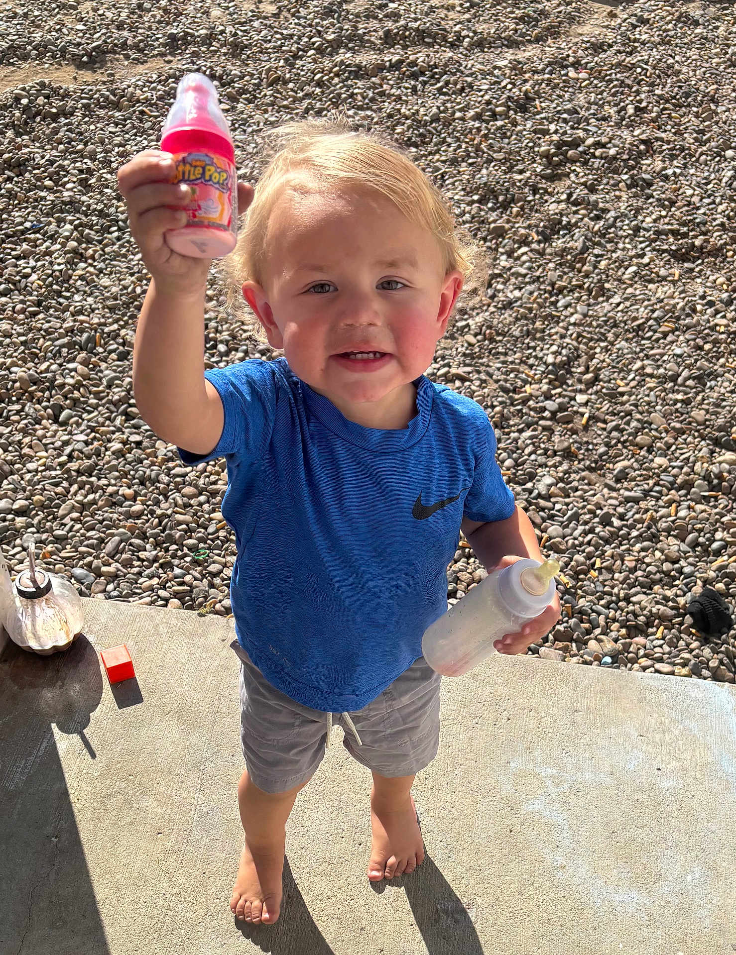 Kyle joined the competition — help win amazing prizes! toddler, child, blue_shirt, barefoot, baby_bottle, pink_bottle, outdoor, gravel, concrete, sunlight, happy, smiling, shorts, person, holding, summer, daylight, playful, young_child, casual_clothing