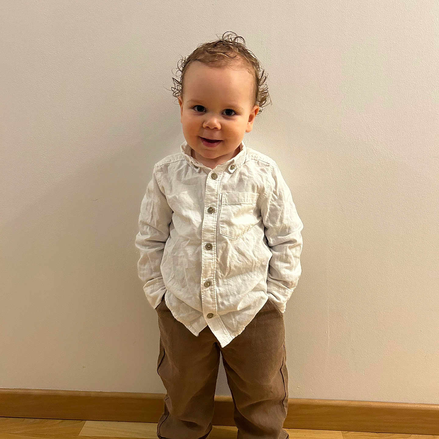 Jules participe au concours pour gagner de l'argent avec cette photo : toddler, child, boy, standing, smiling, curly_hair, white_shirt, brown_pants, socks, indoors, wooden_floor, plain_wall, portrait, cute, hands_in_pockets, happy, casual_clothing, young_child, full_body, light_skinned