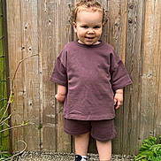 Jules participe au concours pour gagner de l'argent avec cette photo : toddler, child, curly_hair, purple_clothing, shorts, tshirt, sneakers, outdoor, wooden_fence, greenery, plant, standing, smiling, person, casual, daylight, portrait, concrete_ground, cute, young_child