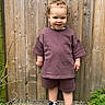 toddler, child, curly_hair, purple_clothing, shorts, tshirt, sneakers, outdoor, wooden_fence, greenery, plant, standing, smiling, person, casual, daylight, portrait, concrete_ground, cute, young_child