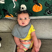Silas is registered to the contest to win money with this photo: baby, child, couch, bib, yellow_shirt, colorful_shorts, sunglasses, gray_couch, green_blanket, skull_pattern, pumpkin_pattern, crow_pattern, mushroom_pattern, indoor, seated, wide_eyes, expression, cute, portrait, cozy