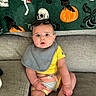 baby, child, couch, bib, yellow_shirt, colorful_shorts, sunglasses, gray_couch, green_blanket, skull_pattern, pumpkin_pattern, crow_pattern, mushroom_pattern, indoor, seated, wide_eyes, expression, cute, portrait, cozy