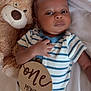 baby, infant, teddy_bear, onesie, striped_clothing, wooden_sign, one_month, bed_sheet, lying_down, hand, face, cute, child, soft_toy, newborn, portrait, sleepwear, resting, indoor, smile