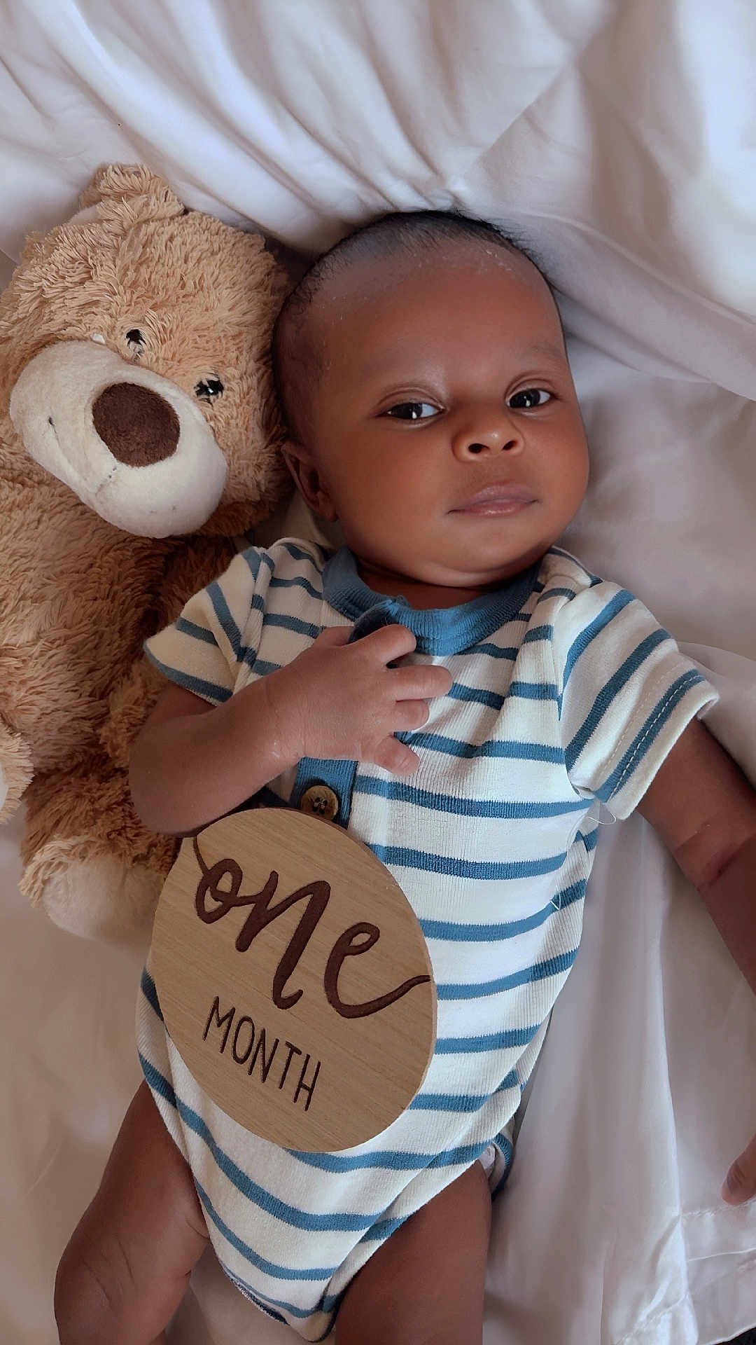 Taige is registered to the contest to win money with this photo: baby, infant, teddy_bear, onesie, striped_clothing, wooden_sign, one_month, bed_sheet, lying_down, hand, face, cute, child, soft_toy, newborn, portrait, sleepwear, resting, indoor, smile
