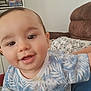 baby, child, close_up, smiling, blue_shirt, patterned_clothing, indoor, living_room, couch, furniture, person, happy, young_child, face, head, curious, sitting, sleeves, home, casual