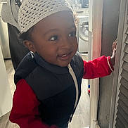 Khessan participe au concours pour gagner de l'argent avec cette photo : toddler, child, hat, knit_hat, vest, red_sweater, smile, indoor, home, flooring, door, hand, curiosity, young_child, face, portrait, clothing, light, window, appliance
