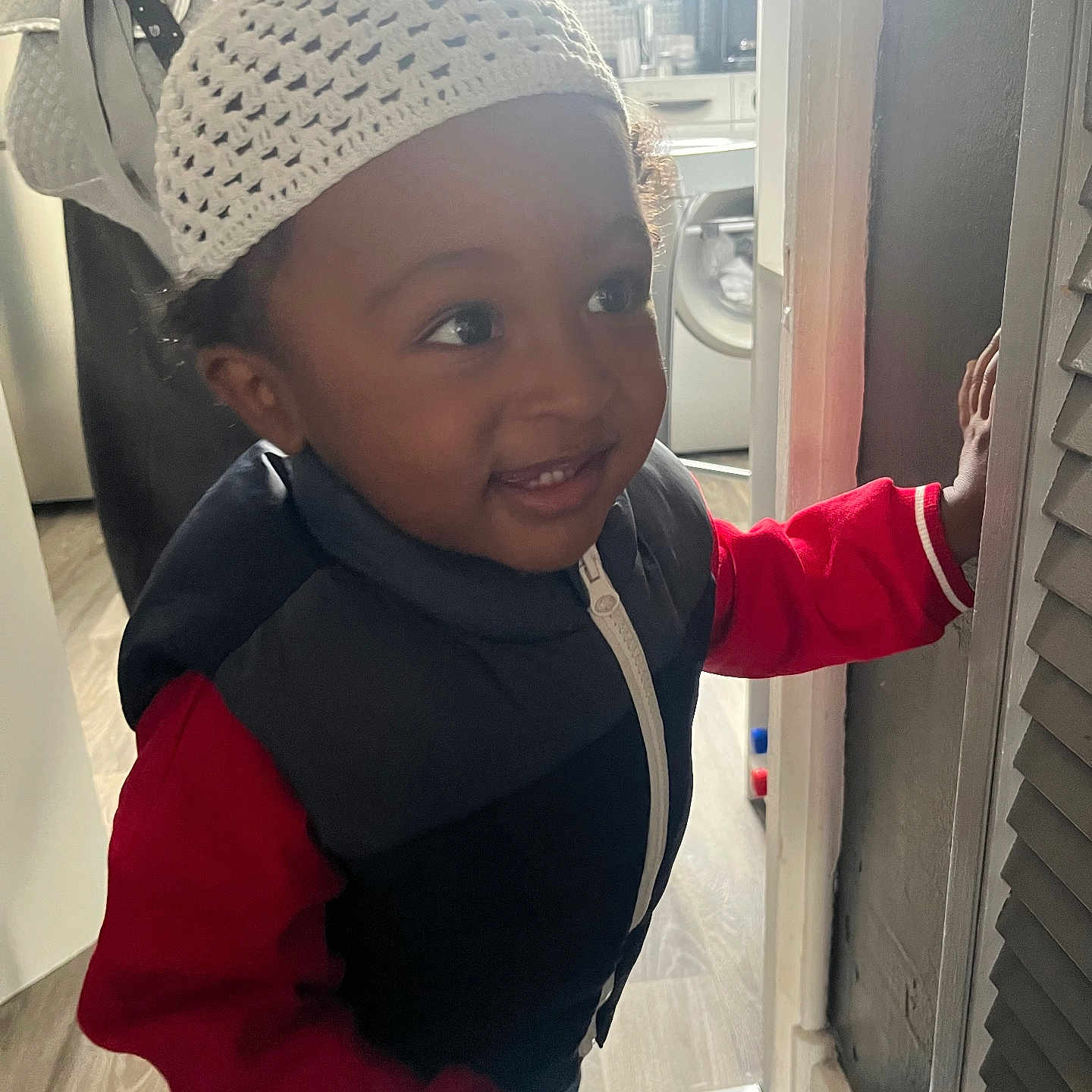 Khessan participe au concours pour gagner de l'argent avec cette photo : appliance, child, clothing, curiosity, door, face, flooring, hand, hat, home, indoor, knit_hat, light, portrait, red_sweater, smile, toddler, vest, window, young_child