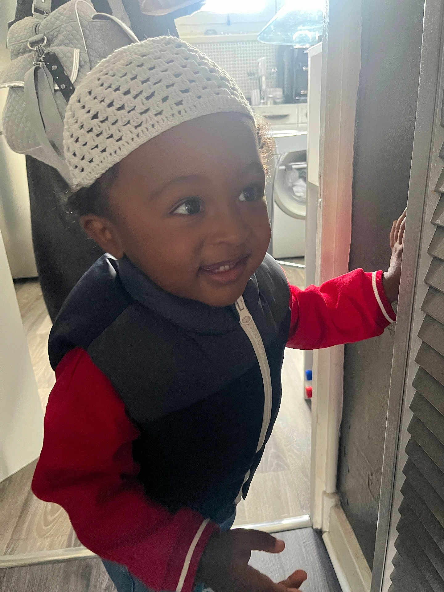 Khessan participe au concours pour gagner de l'argent avec cette photo : toddler, child, hat, knit_hat, vest, red_sweater, smile, indoor, home, flooring, door, hand, curiosity, young_child, face, portrait, clothing, light, window, appliance
