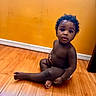 child, toddler, curly_hair, sitting, wooden_floor, orange_wall, indoor, barefoot, diaper, cute, portrait, young_child, skin, baby, expression, looking_up, flooring, home, innocence, casual