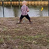 child, grass, water, canal, fence, toy_golf_club, plaid_shirt, boots, outdoor, back_view, young_child, playing, recreation, nature, yard, small_person, casual_clothing, daytime, activity, lawn