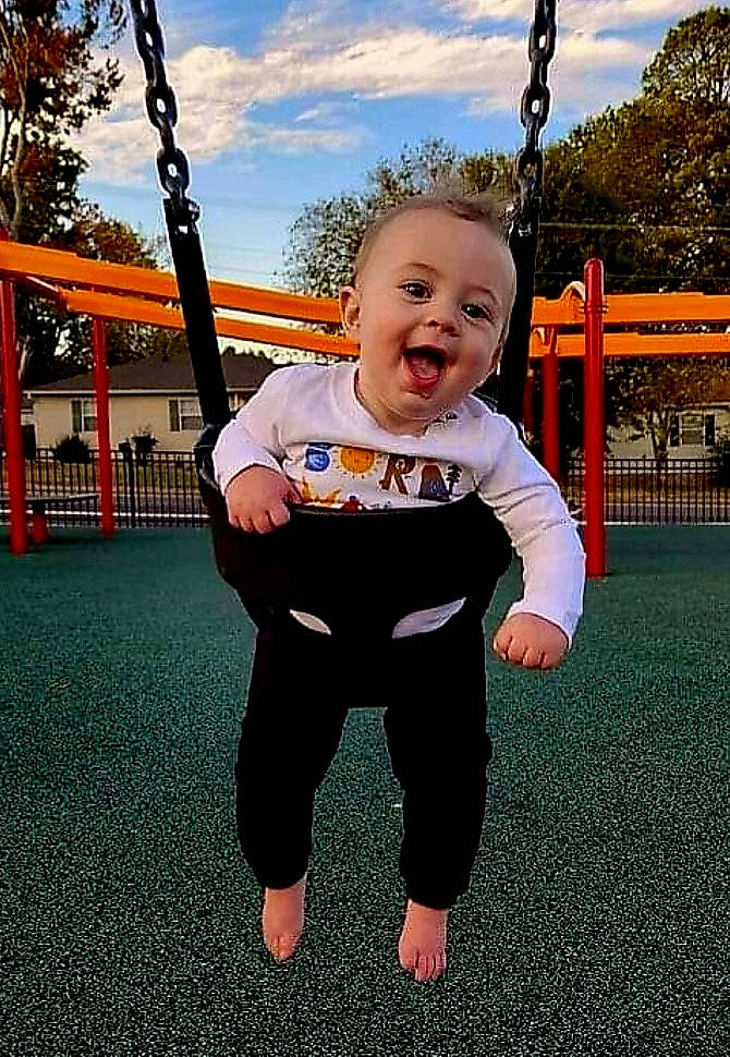 Callum is registered to the contest to win money with this photo: baby, baby_toddler_clothing, city, cloud, facial_expression, fun, grass, happy, leisure, outdoor_play_equipment, people_in_nature, person, plant, playground, recreation, sky, sleeve, smile, swing, toddler