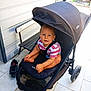 Nhaël a rejoint le concours — aidez-le/la à gagner de superbes lots ! toddler, child, stroller, outdoor, bench, footwear, pavement, smiling, curly_hair, sunlight, clothing, striped_shirt, baby, seat, wheels, black_stroller, happy, person, daylight, casual