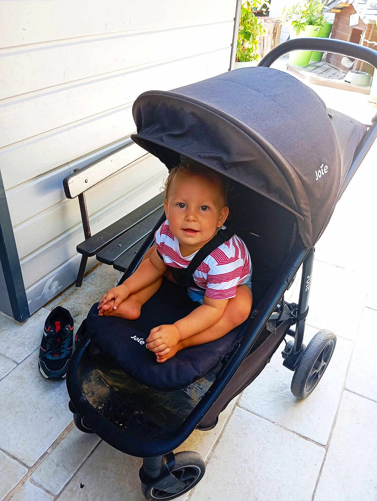 Nhaël a rejoint le concours — aidez-le/la à gagner de superbes lots ! toddler, child, stroller, outdoor, bench, footwear, pavement, smiling, curly_hair, sunlight, clothing, striped_shirt, baby, seat, wheels, black_stroller, happy, person, daylight, casual