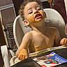 child, toddler, baby, high_chair, eating, food, messy_face, orange_food, portrait, sitting, indoors, home, dining, table, tray, bare_chested, curly_hair, smiling, facial_expression, family_scene