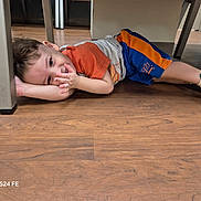 Ezra joined the competition — help win amazing prizes! child, boy, toddler, lying_down, indoor, floor, wooden_floor, table, furniture, playful, casual_clothing, orange_shirt, blue_shorts, sneakers, colorful_shoes, smiling, face, portrait, home, photograph