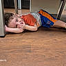 child, boy, toddler, lying_down, indoor, floor, wooden_floor, table, furniture, playful, casual_clothing, orange_shirt, blue_shorts, sneakers, colorful_shoes, smiling, face, portrait, home, photograph