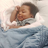 Ember is registered to the contest to win money with this photo: baby, infant, smile, sleepy, curly_hair, blanket, pink_blanket, blue_blanket, bed, pillow, plush_toy, stuffed_animal, bedding, clothing, gray_shirt, lying_down, cute, portrait, closeup, nursery