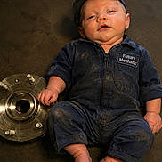 Atlas is registered to the contest to win money with this photo: baby, infant, child, portrait, face, serious_expression, cap, coveralls, jumpsuit, mechanic, workshop, metal_part, machinery, garage, industrial, floor, sitting, bare_feet, fashion, photography