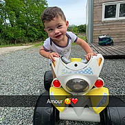 Kaïs participe au concours pour gagner de l'argent avec cette photo : child, toddler, toy_vehicle, outdoor, gravel, greenery, shed, smiling, happy, casual, daylight, short_hair, playing, front_view, baby, young_child, vehicle, nature, grass, cute