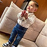 toddler, child, standing, indoor, jeans, shirt, sneakers, couch, pillow, wooden_door, floor, person, young_child, curious, casual_clothing, home_interior, fashion, portrait, cute, expression