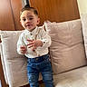 toddler, child, jeans, shirt, gold_chain, sneakers, couch, indoor, portrait, standing, furniture, wooden_background, fashion, cute, person, young_child, casual, home, smiling, accessory