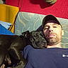 man, dog, black_dog, couch, red_wall, shoe, cap, facial_hair, relaxing, indoor, snuggling, blue_shirt, resting, pet, companion, casual, home, comfort, closeup, portrait
