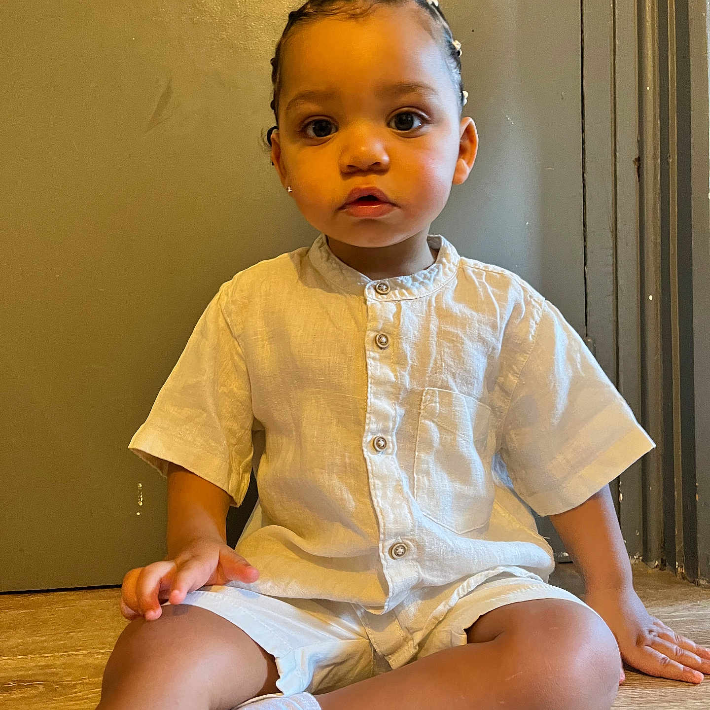 Lyenzo a rejoint le concours — aidez-le/la à gagner de superbes lots ! baby_socks, braided_hair, brown_skin, button_up_shirt, casual_clothing, child, curious, cute, expression, face, floor, indoor, looking_at_camera, portrait, shorts, sitting, toddler, white_shirt, wooden_floor, young_child
