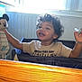 child, toddler, curly_hair, smiling, indoor, playpen, toy, mickey_mouse, pacifier, window, daylight, shirt, happy, furniture, wooden_table, person, face, playing, casual_clothing, home