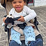 child, toddler, stroller, curly_hair, smiling, plush_toy, mickey_mouse, pants, shoes, outdoor, cobblestone, seatbelt, happy, person, clothing, footwear, toy, casual_wear, daylight, baby